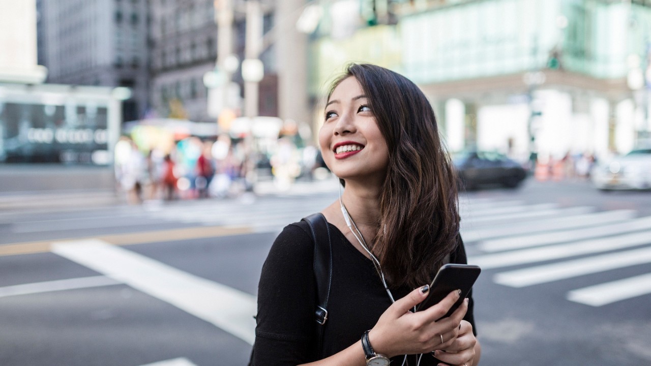 Wanita yang sedang tersenyum menyeberang jalan di kota besar, dengan ponsel pintar di tangan dan earphone di telinganya.