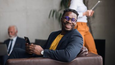 Happy young black man using a phone. Colleagues in the background. Joyful African-American businessman in a suit, sitting in a lobby, holding a phone .