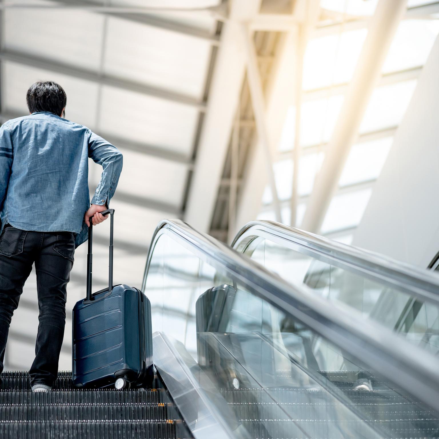 Asian man tourist carrying suitcase luggage and digital tablet on escalator in airport terminal.