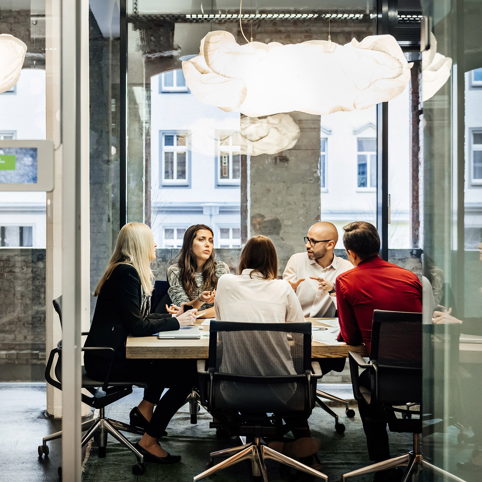 Un grupo de personas está sentado en una mesa durante una reunión de negocios en una oficina moderna y bien iluminada. El equipo está hablando de negocios, mientras que se pueden ver gráficos circulares colgando en la pared. Las grandes ventanas brillantes se ven en segundo plano.