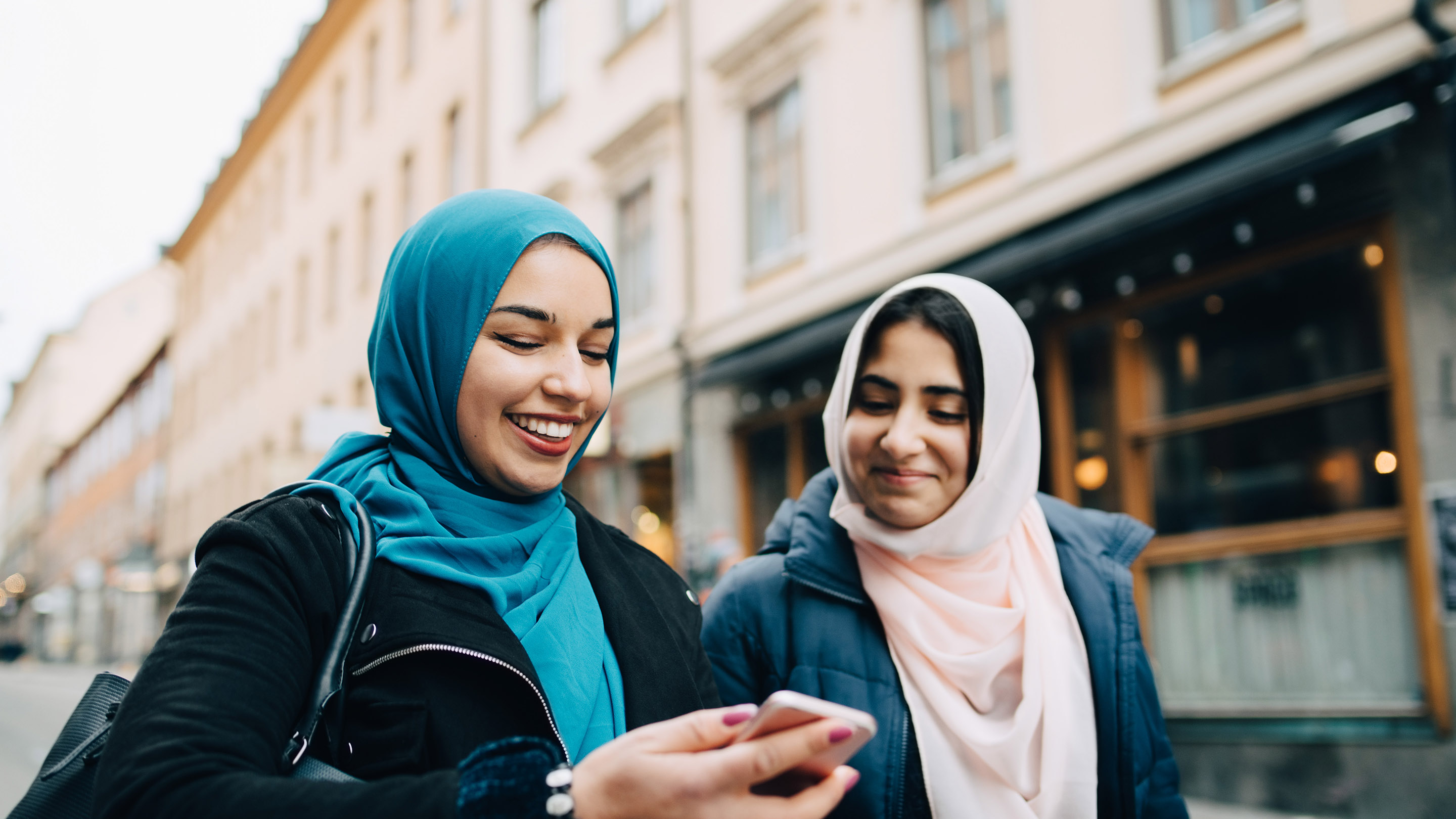 Smiling young woman sharing mobile phone with female friend walking in city