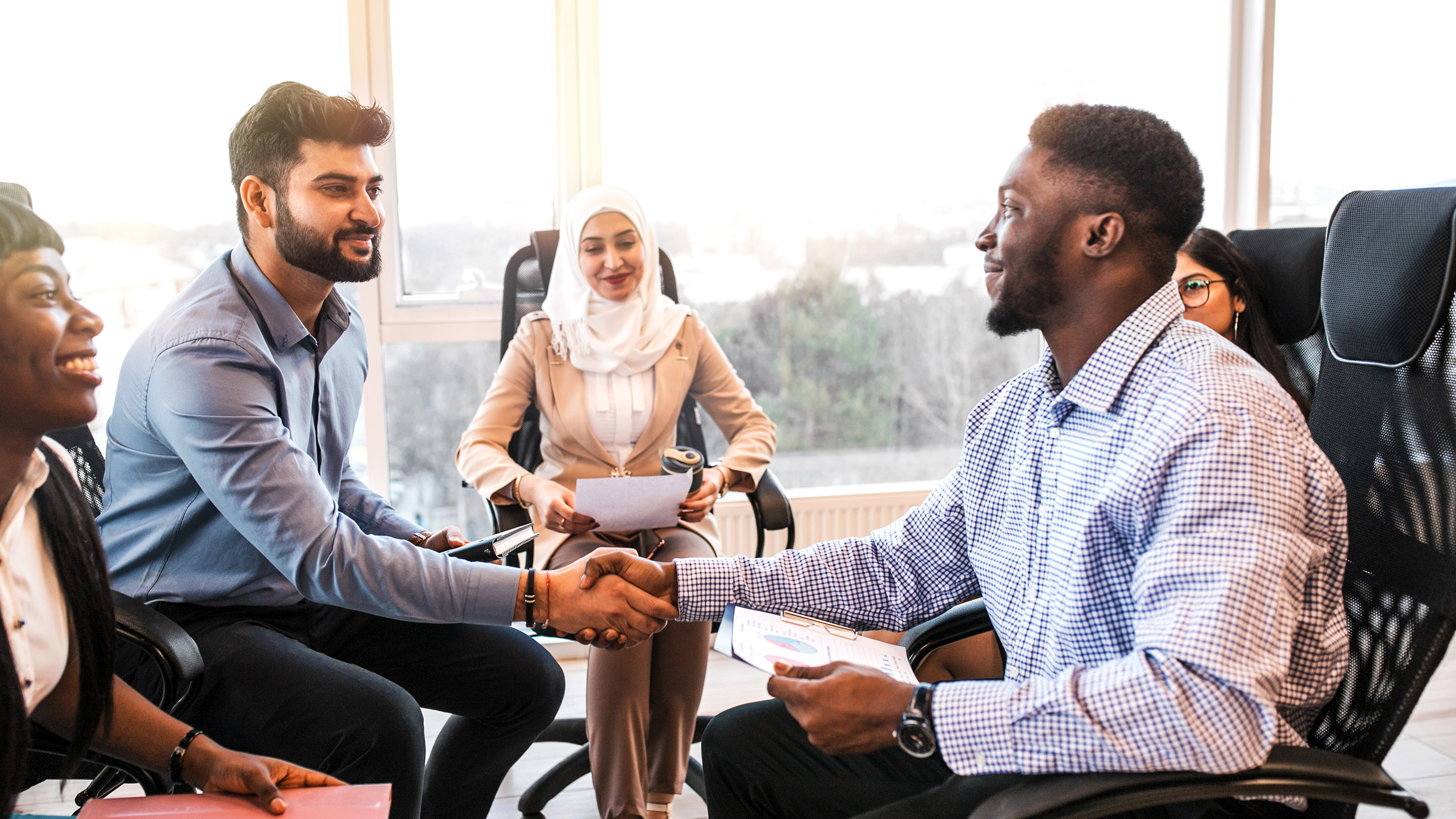Smiling black and Indian businessmen handshaking after signing contract at multi-ethnic meeting with businesswoman.