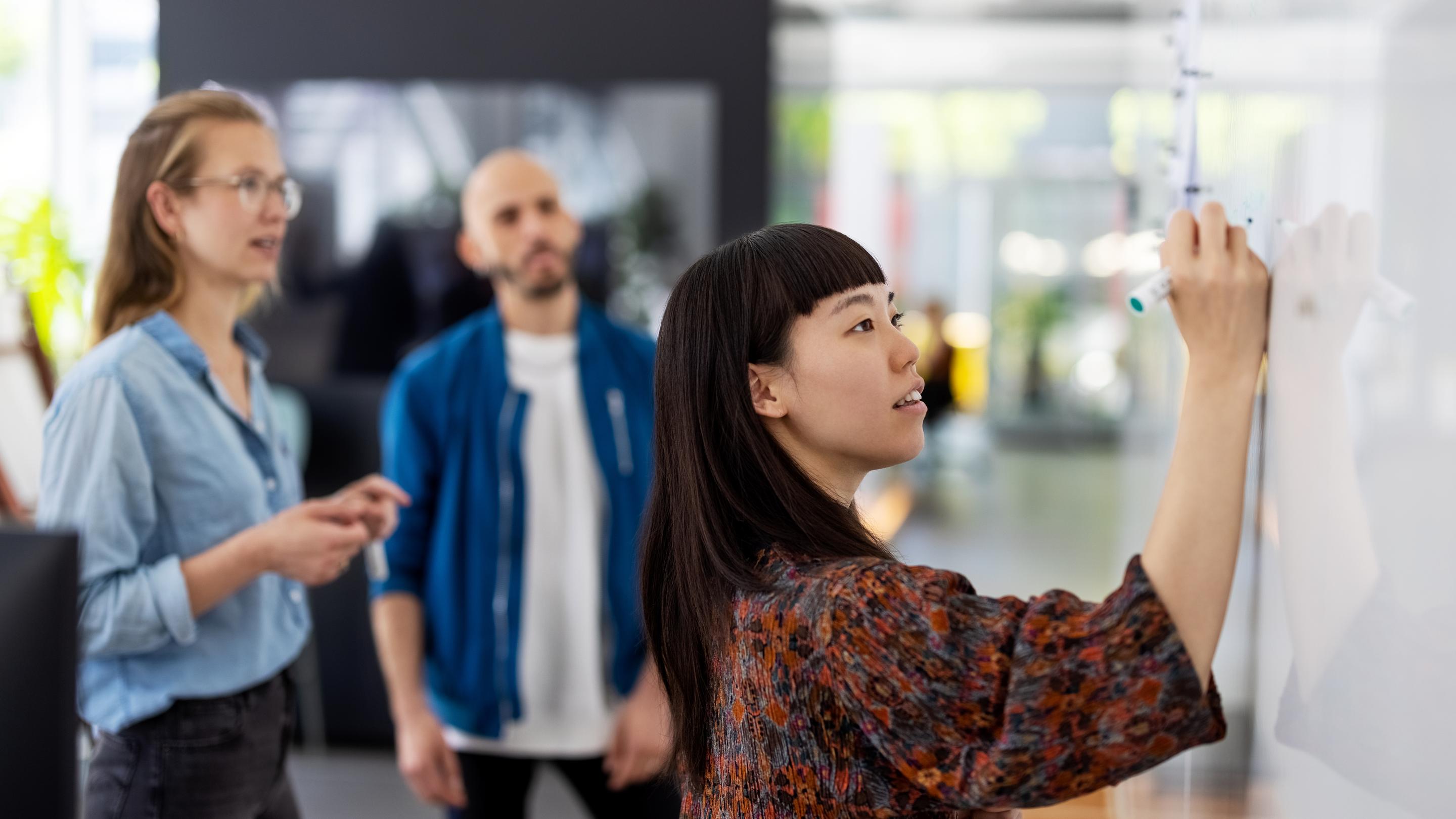 Young businesswoman explaining a business plan to colleague in office. Woman writing on the whiteboard during presentation in hybrid office space.