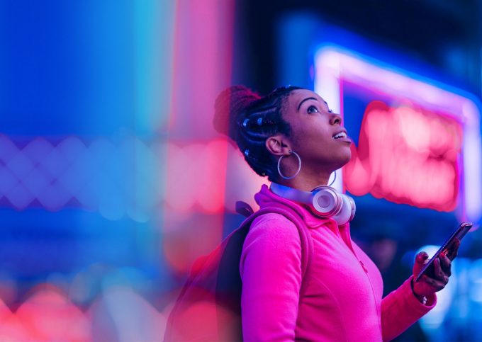 Young Black woman in pink shirt wearing headphones using a smart phone at night with neon lights around her