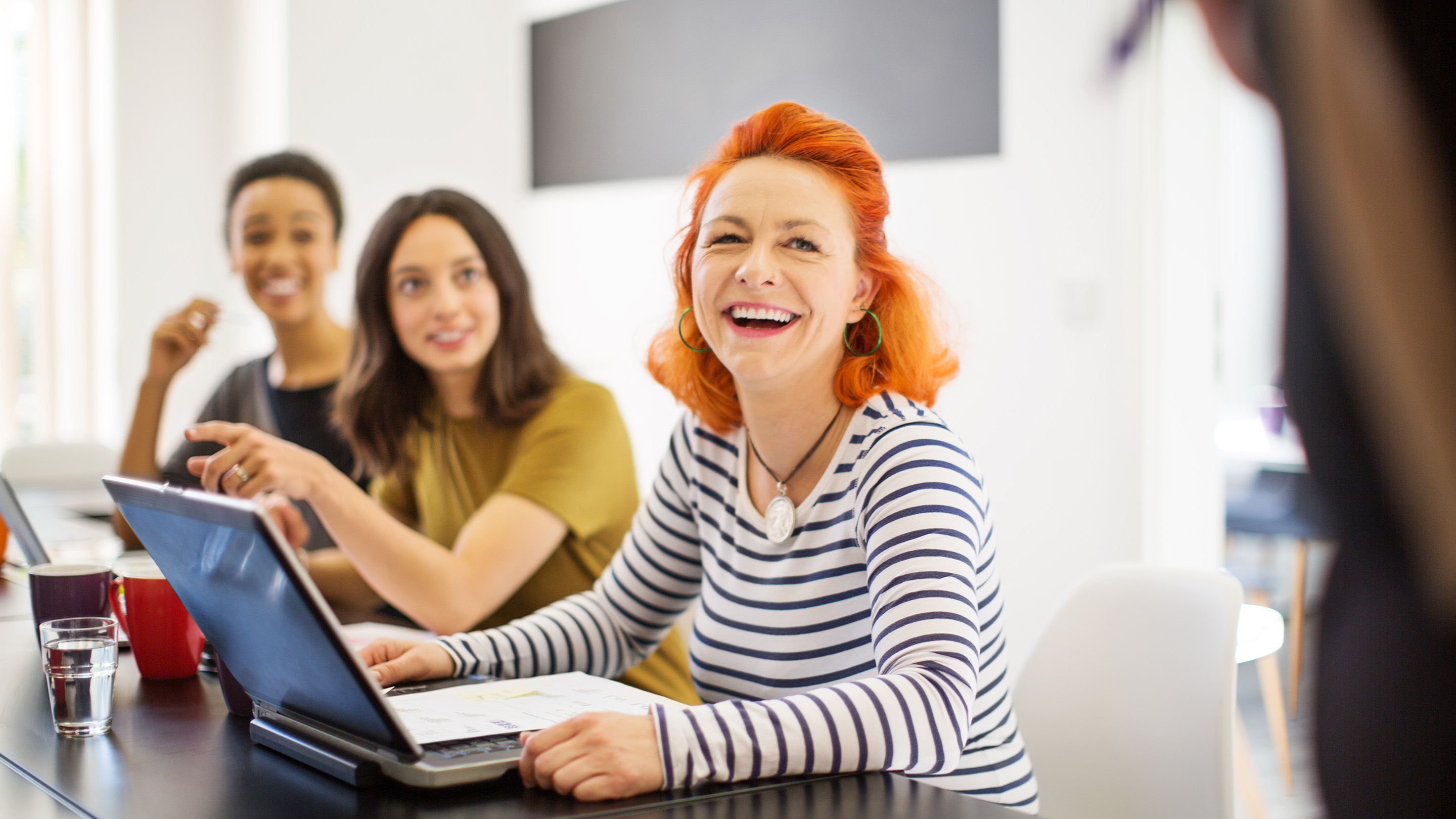 Mature woman smiling in meeting with colleagues in boardroom. Multi-ethnic professionals are sitting at conference table.