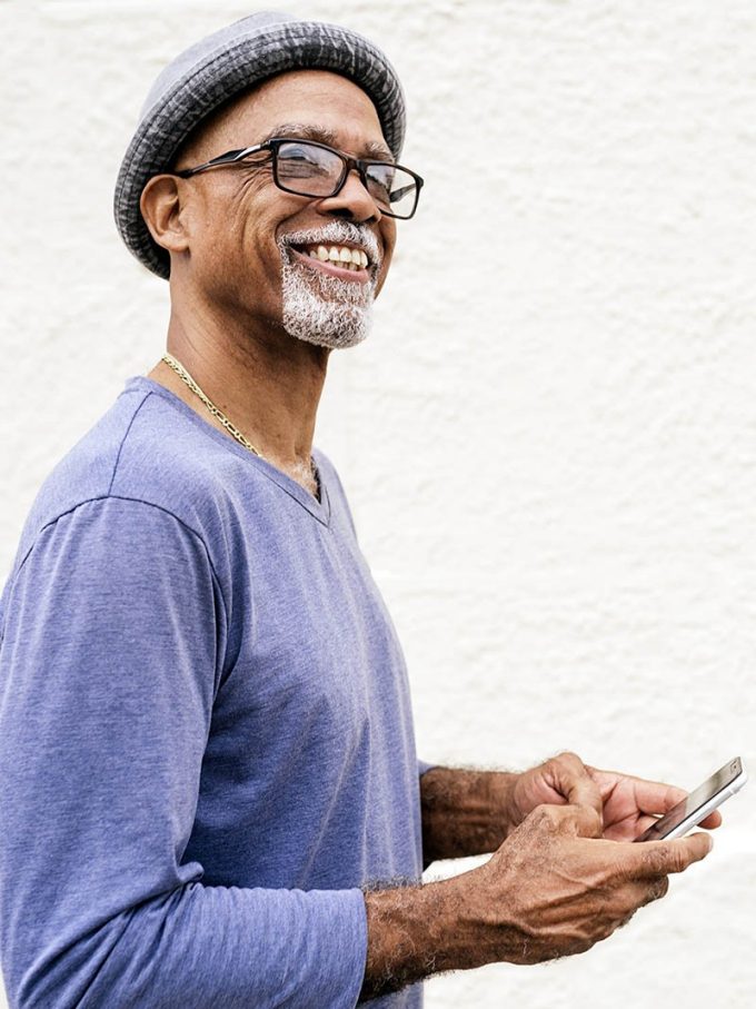 Stock photo of mature afro man standing in front of a white wall. He is looking away from the camera. He is wearing glasses and a hat.