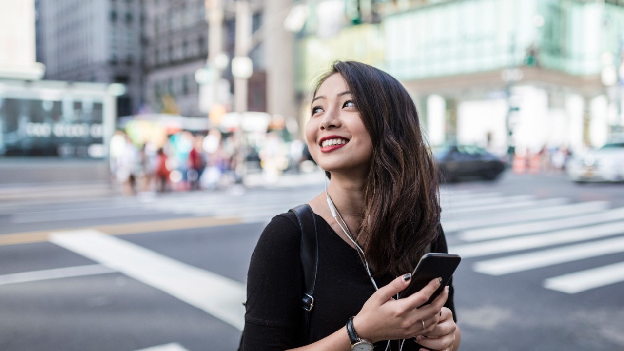 Smilende kvinde, der krydser gaden i en storby med en smartphone i hånden og ørepropper i ørerne.