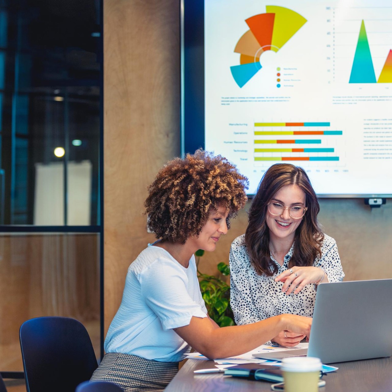 Junge Geschäftskollegen, die in einem modernen Büro an einem Laptop zusammenarbeiten, mit Charts auf einem Fernseher und Papierkram.