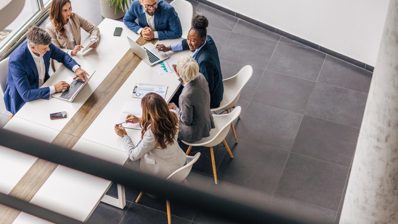 Eine Gruppe Menschen in Business Kleidung sitzt an einem Ende eines Konferenztisches und unterhält sich angeregt. Auf dem Tisch liegen zwei Laptops, Handys und ein paar ausgedruckte Charts. 