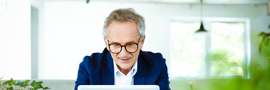 Elderly elegant man wearing navy blue jacket, white shirt and glasses sitting on sofa in the eco-friendly office. Senior businessman having video conference on laptop.