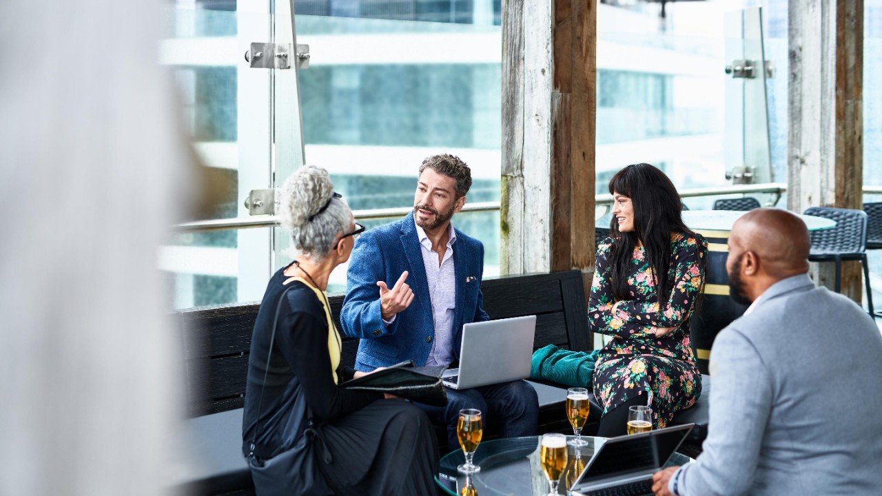 Vier Personen sitzen in einem modernen Raum um einen Tisch mit Getränken und Laptops, im Gespräch vertieft.