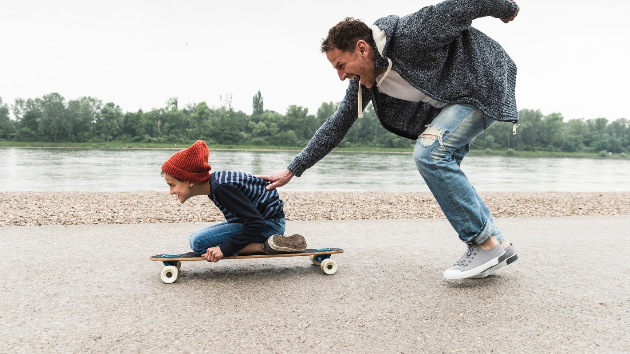 Happy father pushing son on skateboard at the riverside