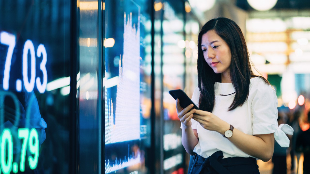 Confidence young Asian businesswoman checking financial trading data on smartphone by the stock exchange market display screen board in downtown financial district