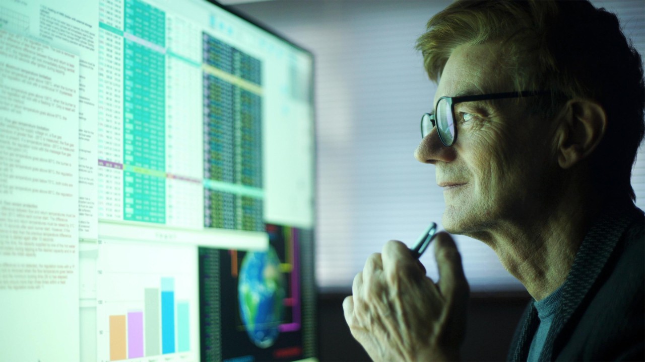 Stock photo of a mature man working in his home office. He’s studying a large computer monitor displaying a variety of numerical data, global information & text.