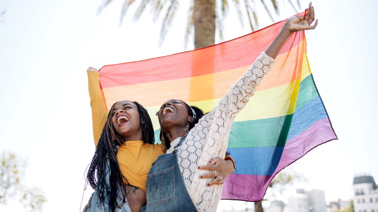 Low angle of delighted African American females in love hugging on street while standing with LGBT rainbow flag and looking away