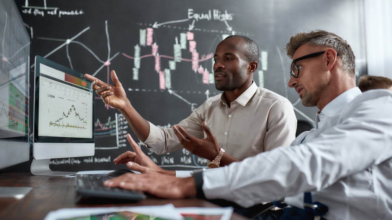 Two diverse colleagues traders talking to each other, looking at graphs while sitting in the office in front of multiple computer screens. Stock trading, people, business concept.