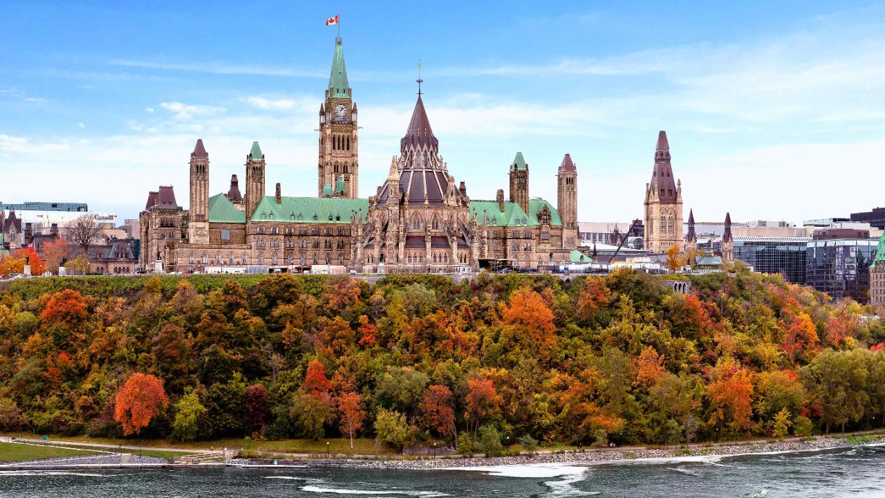 Parliament Hill showcases stunning autumn colors as vibrant leaves contrast with the historic architecture. The Ottawa River flows gently nearby, enhancing the picturesque landscape.