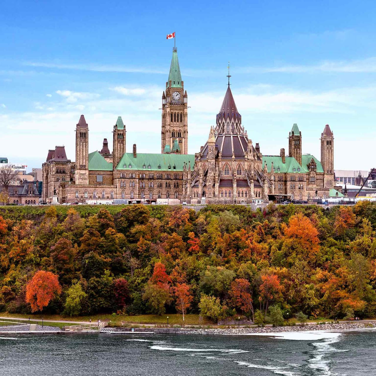 Parliament Hill showcases stunning autumn colors as vibrant leaves contrast with the historic architecture. The Ottawa River flows gently nearby, enhancing the picturesque landscape.