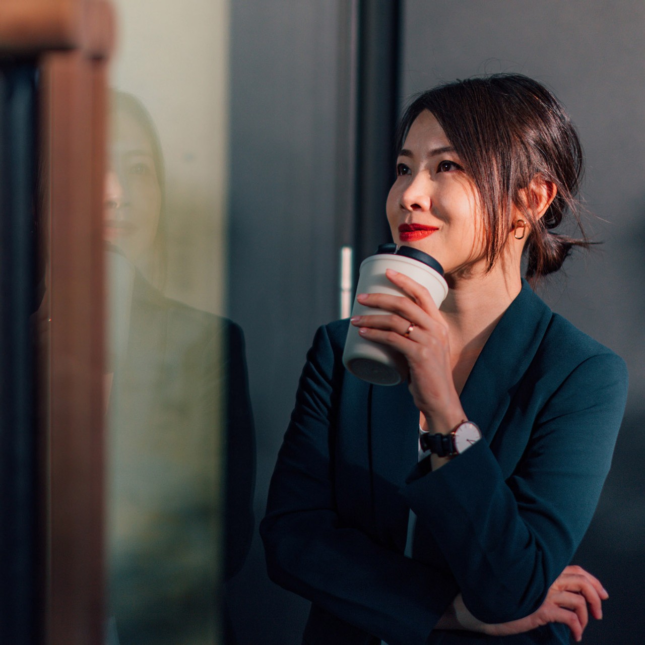 Confident Asian woman looking out the window, holding a reusable coffee cup, determined to succeed.