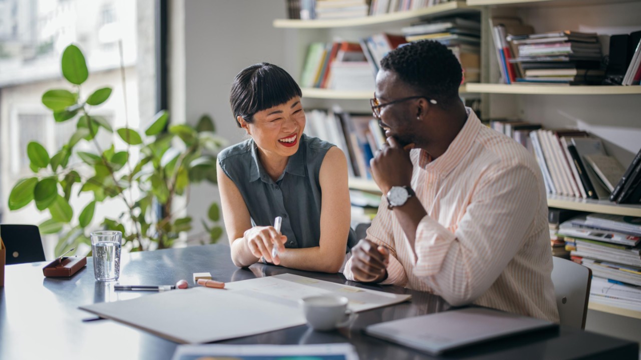 A laughing Japanese businesswoman having a conversation with her African-American coworker while they are sitting at the desk.