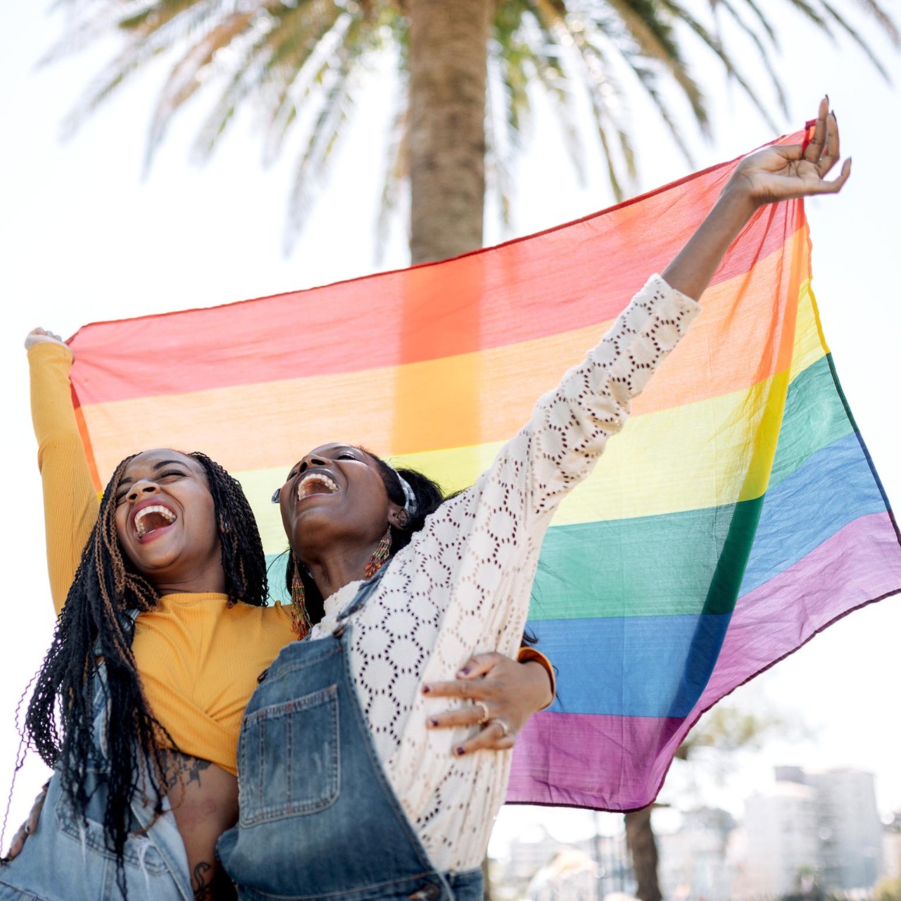 Low angle of delighted African American females in love hugging on street while standing with LGBT rainbow flag and looking away