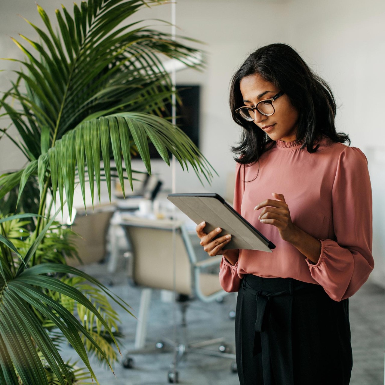 Female executive with medium-length black hair wearing eyeglasses and businesswear standing outside modern meeting room and checking data on tablet.