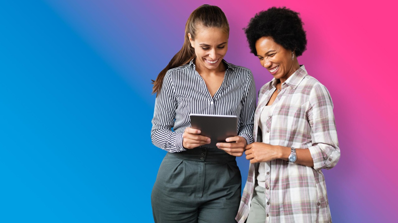 two women on blue to pink gradient background smiling and looking at tablet