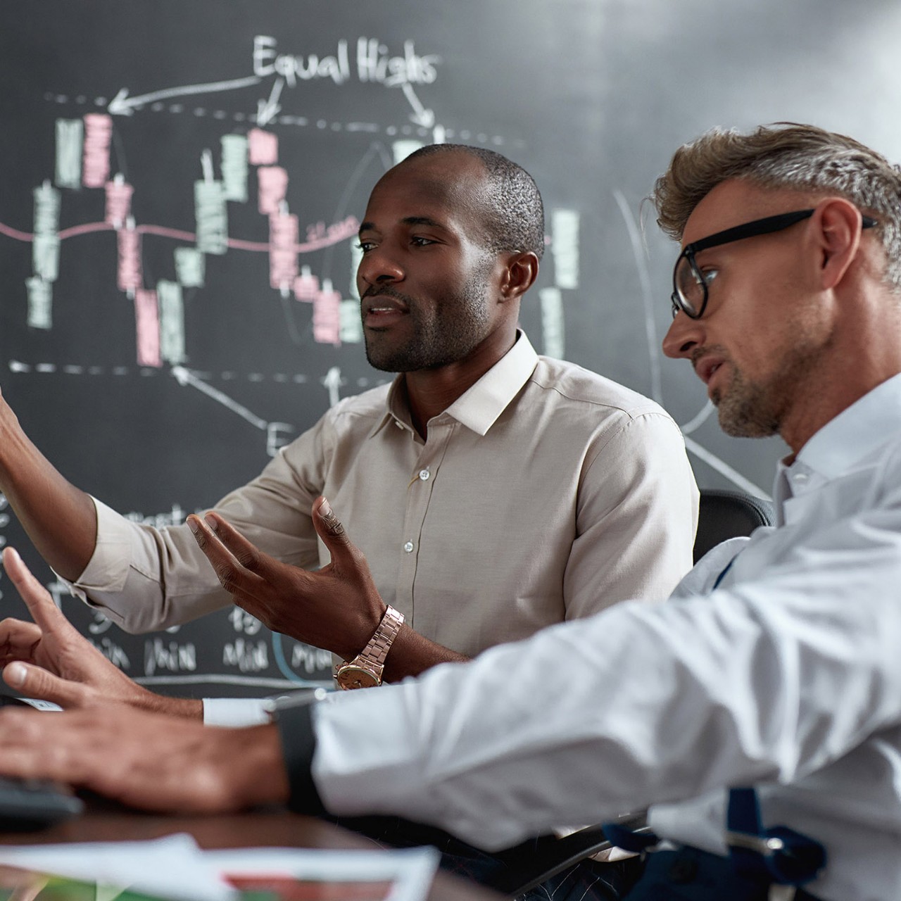 Two diverse colleagues traders talking to each other, looking at graphs while sitting in the office in front of multiple computer screens. Stock trading, people, business concept.