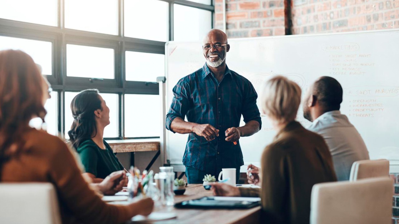 A senior man leads a discussion in a well-lit conference room filled with professionals.