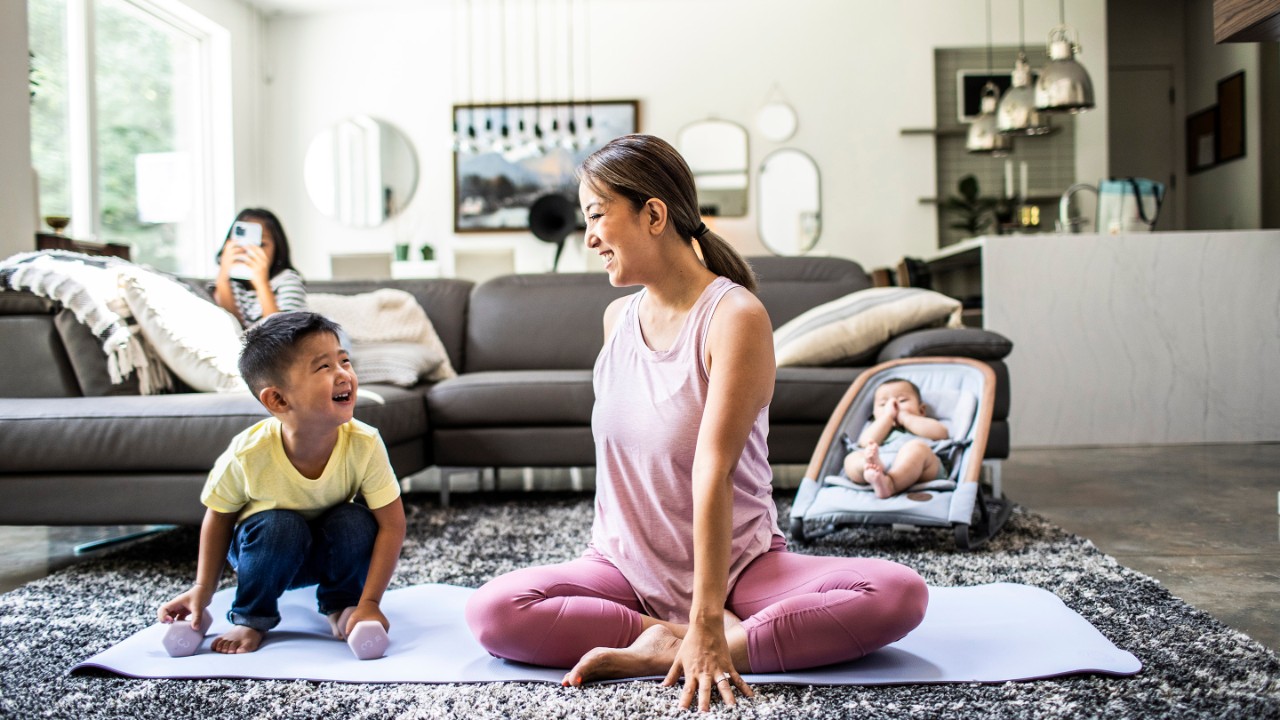 Woman sitting on yoga mat in living room, with young child next to her, both smiling and happy, baby in background and young girl sitting on sofa