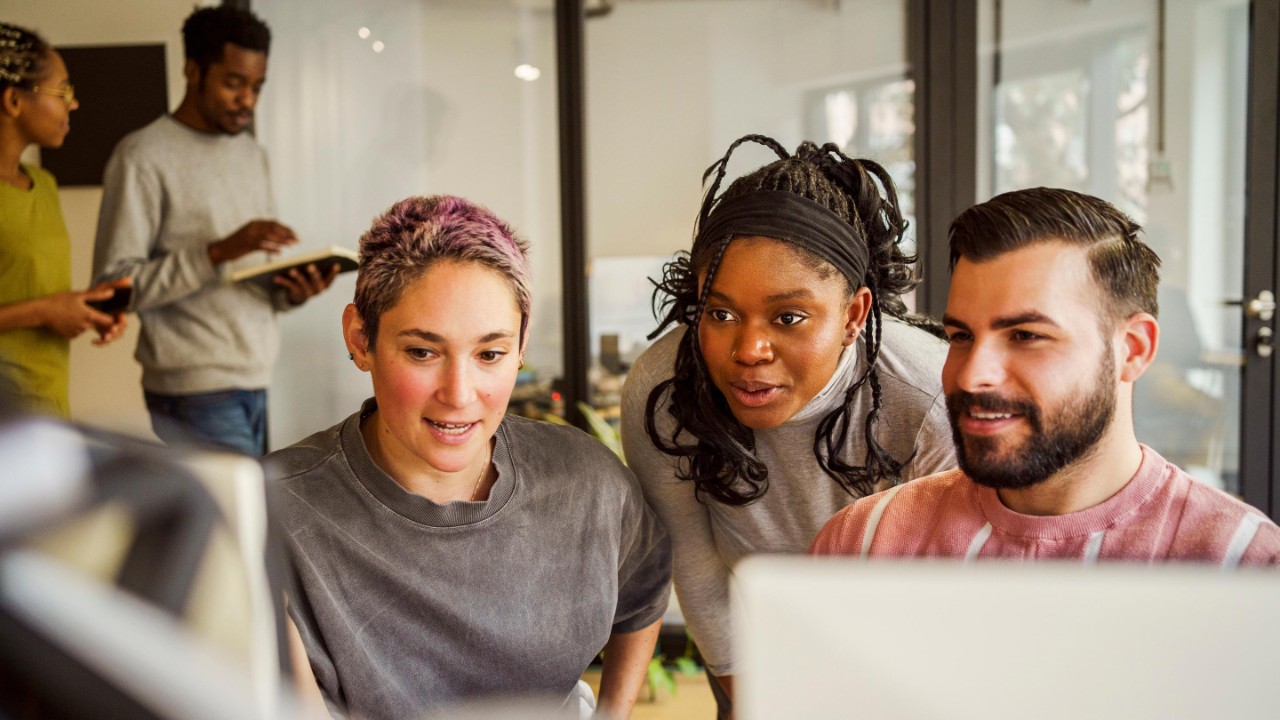 Group of young office workers at a computer screen, man and woman walk past in the background