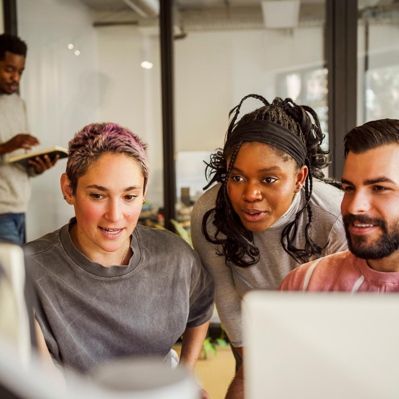 Group of young office workers at a computer screen, man and woman walk past in the background