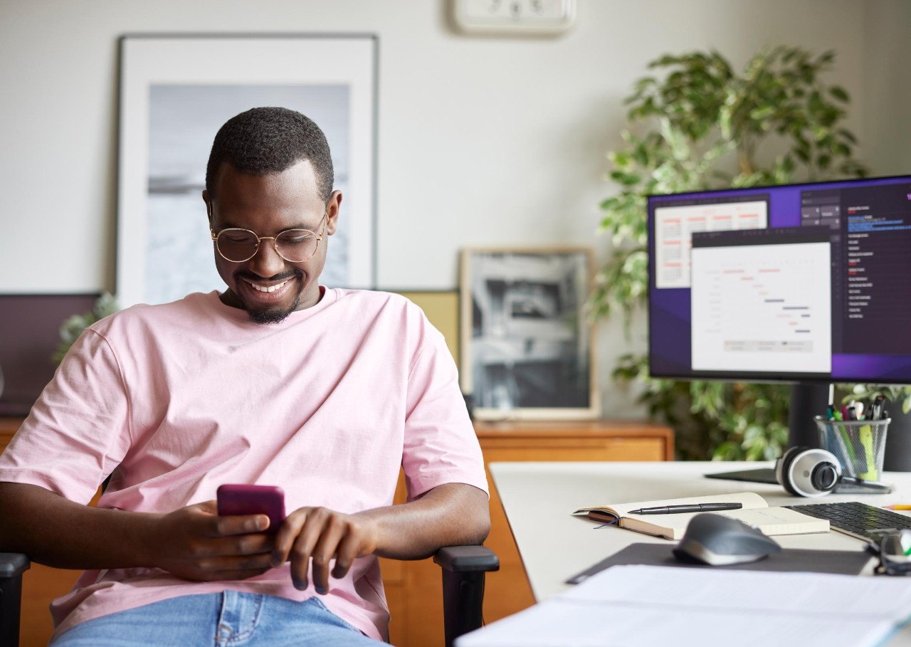 Smiling businessman using smart phone while sitting by computer desk. Male professional is surfing internet at home office. He is in casuals at apartment.