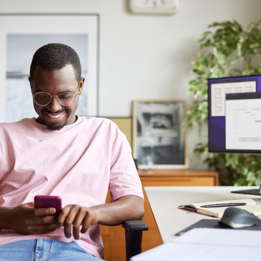 Smiling businessman using smart phone while sitting by computer desk. Male professional is surfing internet at home office. He is in casuals at apartment.