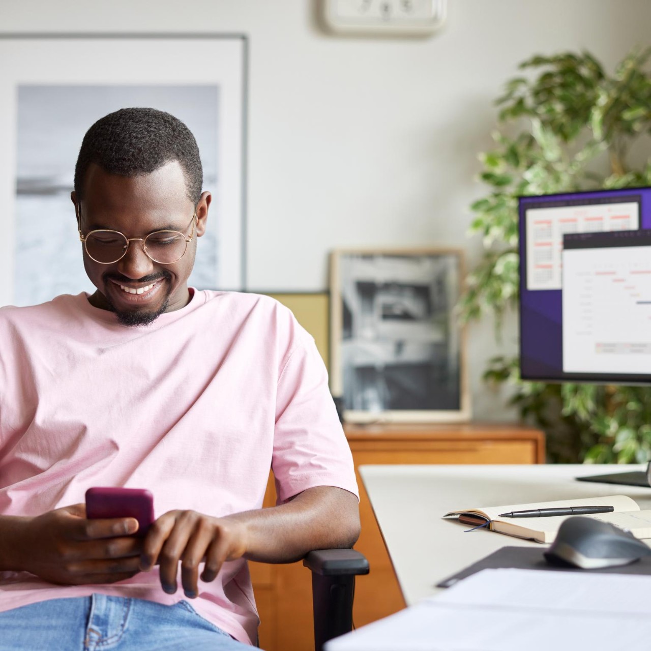 Smiling businessman using smart phone while sitting by computer desk. Male professional is surfing internet at home office. He is in casuals at apartment.