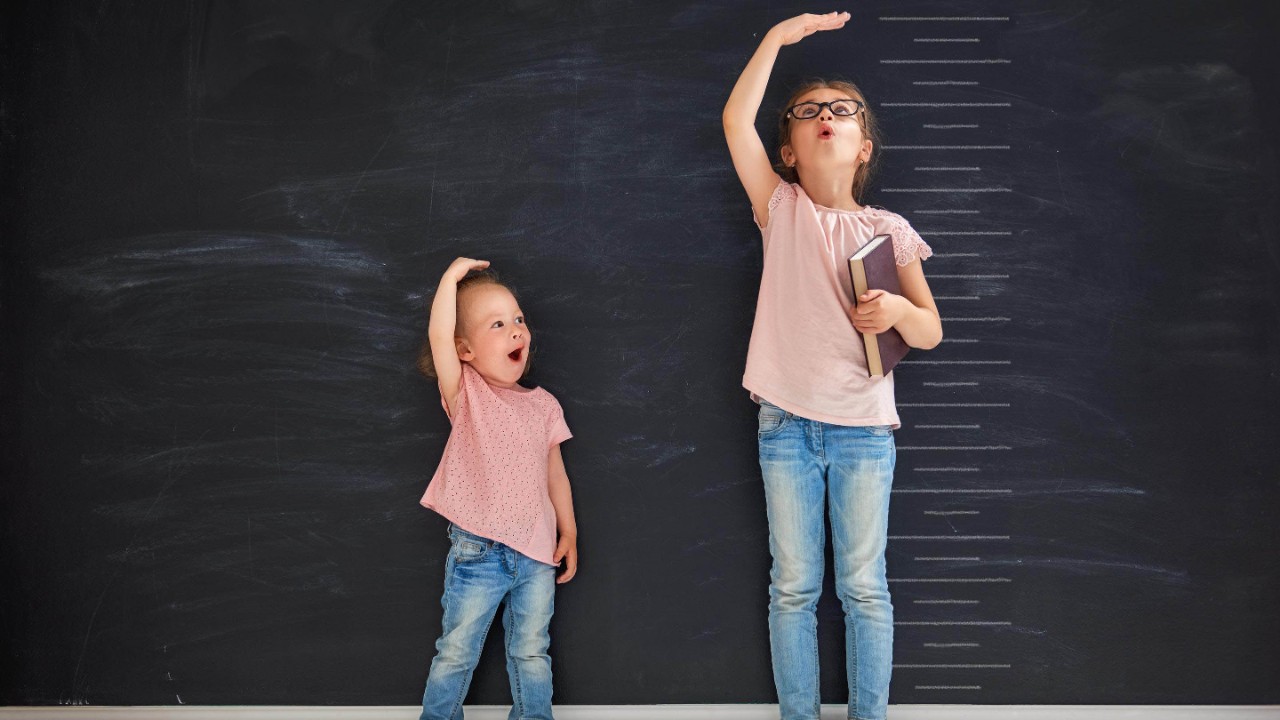 Two children sisters play together. Kid measures the growth on the background of blackboard. Concept of education.