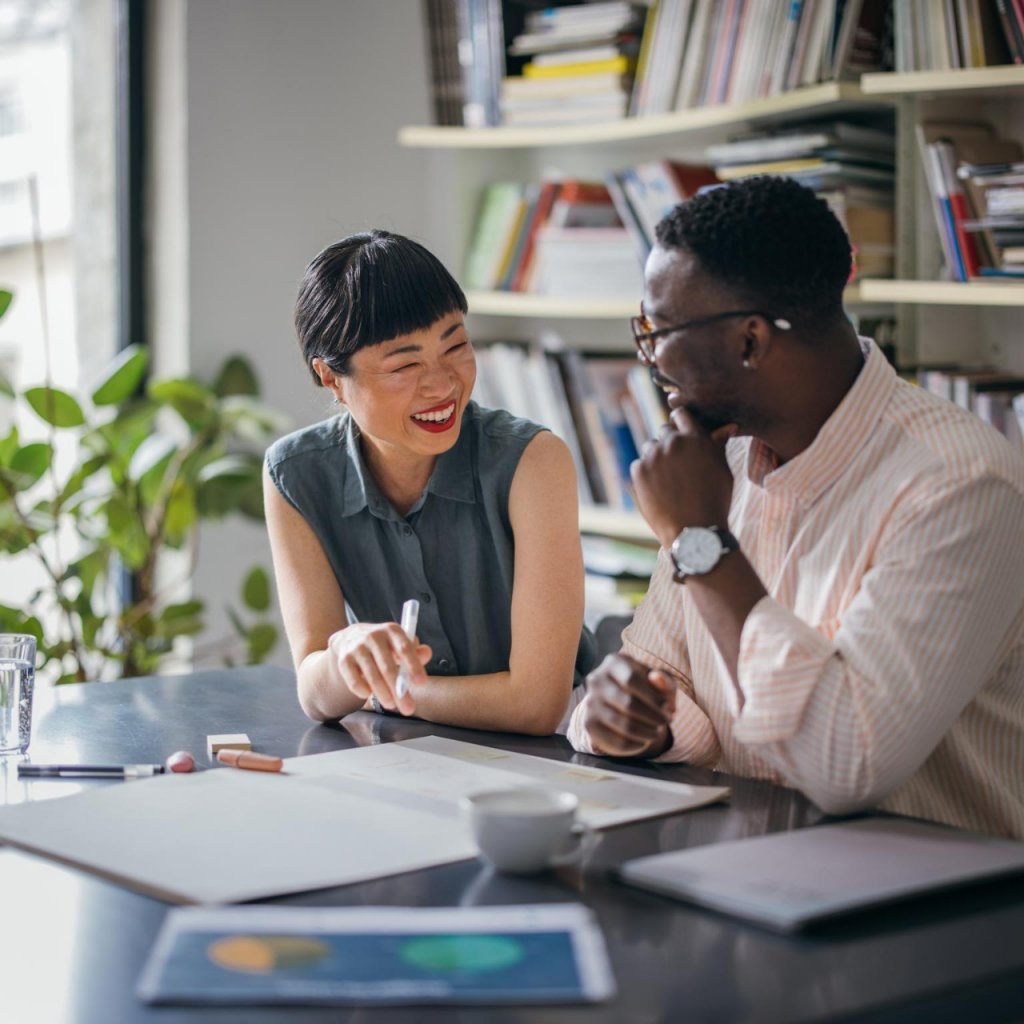 A laughing Japanese businesswoman having a conversation with her African-American coworker while they are sitting at the desk.