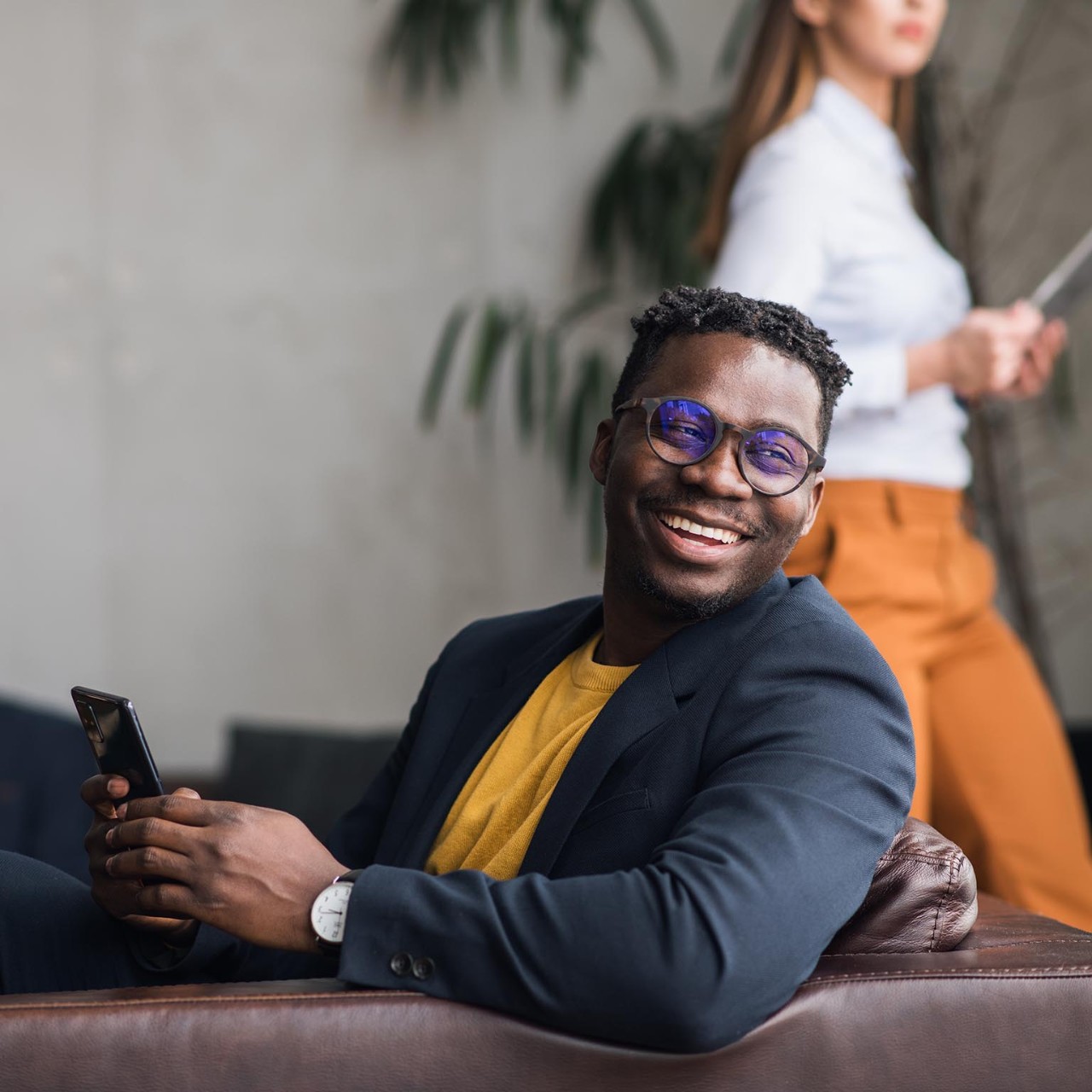 Happy young black man using a phone. Colleagues in the background. Joyful African-American businessman in a suit, sitting in a lobby, holding a phone .