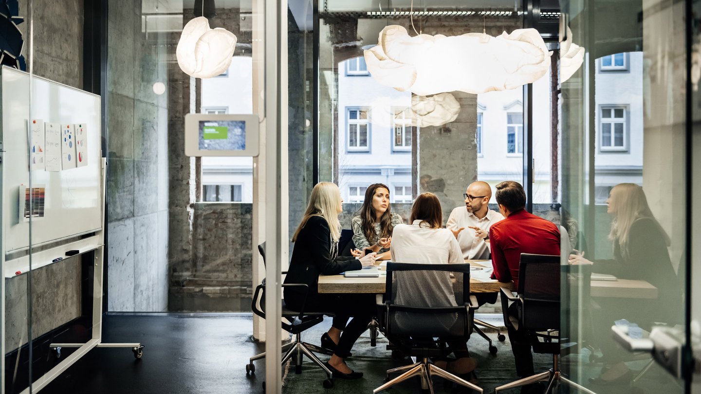  A group of people is sitting at a table during a business meeting in a bright, modern office. The team is talking business while pie charts can be seen hanging on the wall. Big bright windows are seen in the background.