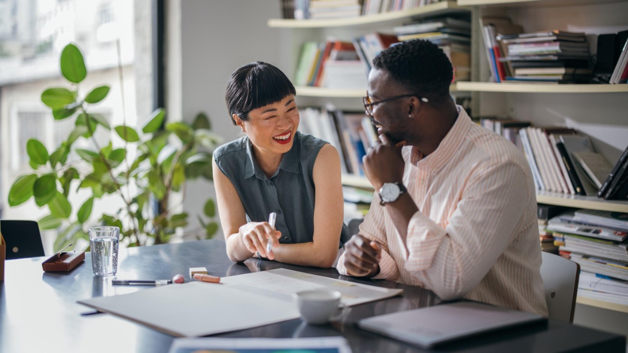 A laughing Japanese businesswoman having a conversation with her African-American coworker while they are sitting at the desk.