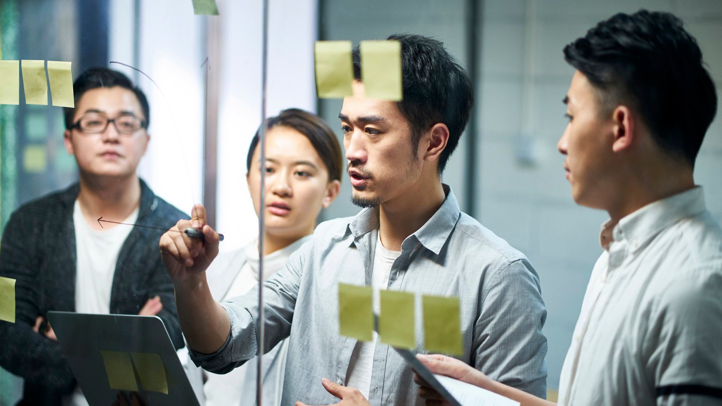 young asian entrepreneur of small company drawing a diagram on glass during team meeting discussing and analysing business situation in office.