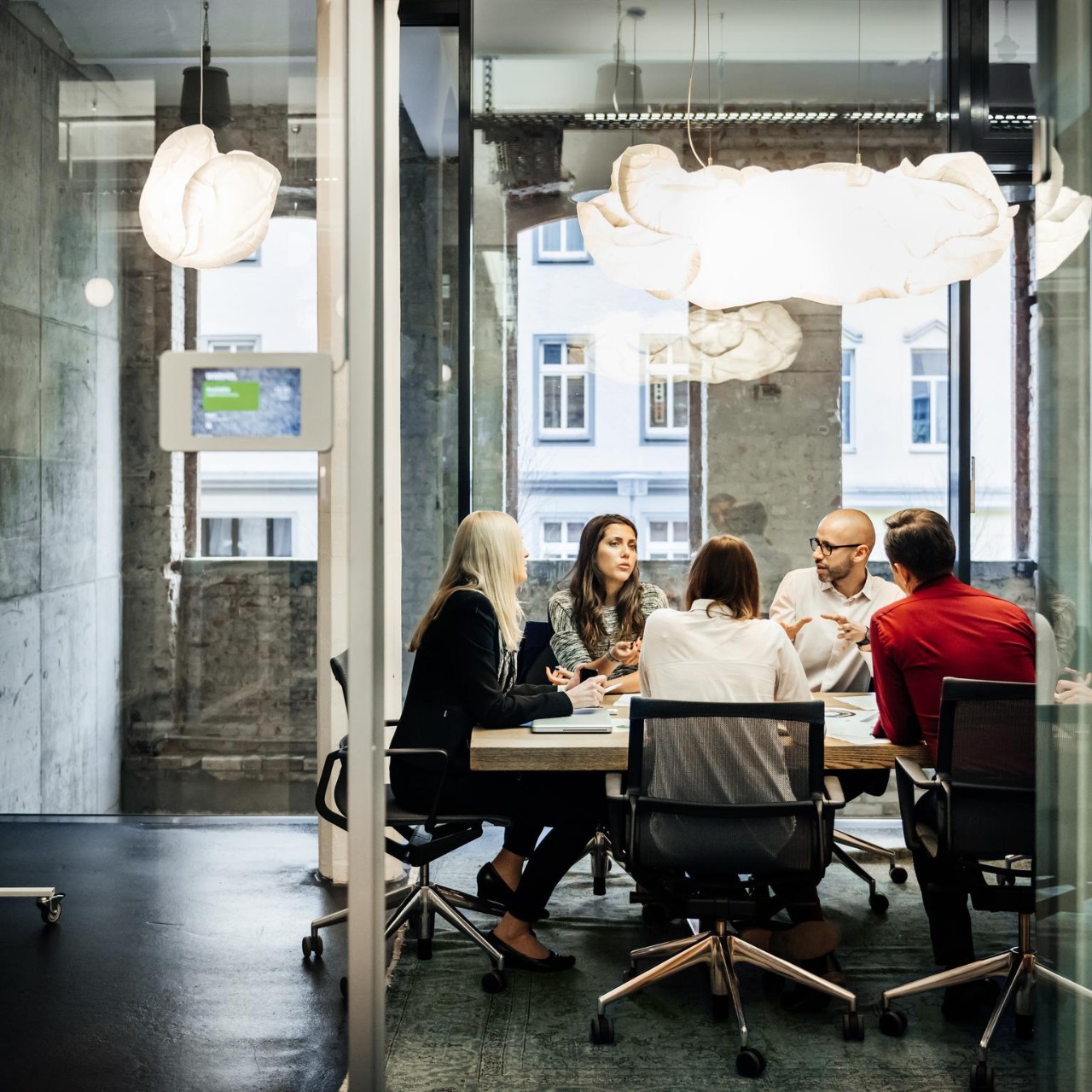A group of people is sitting at a table during a business meeting in a bright, modern office. The team is talking business while pie charts can be seen hanging on the wall. Big bright windows are seen in the background.