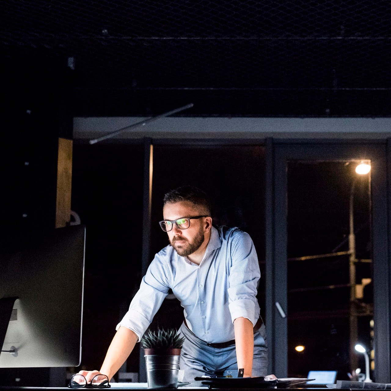 Thoughtful hipster businessman in his office working late at night at desk with computer and laptop.