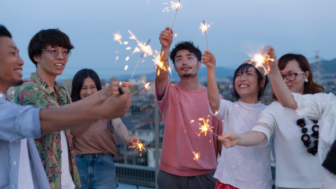 Group of young Japanese men and women with sparklers on a rooftop in an urban setting.