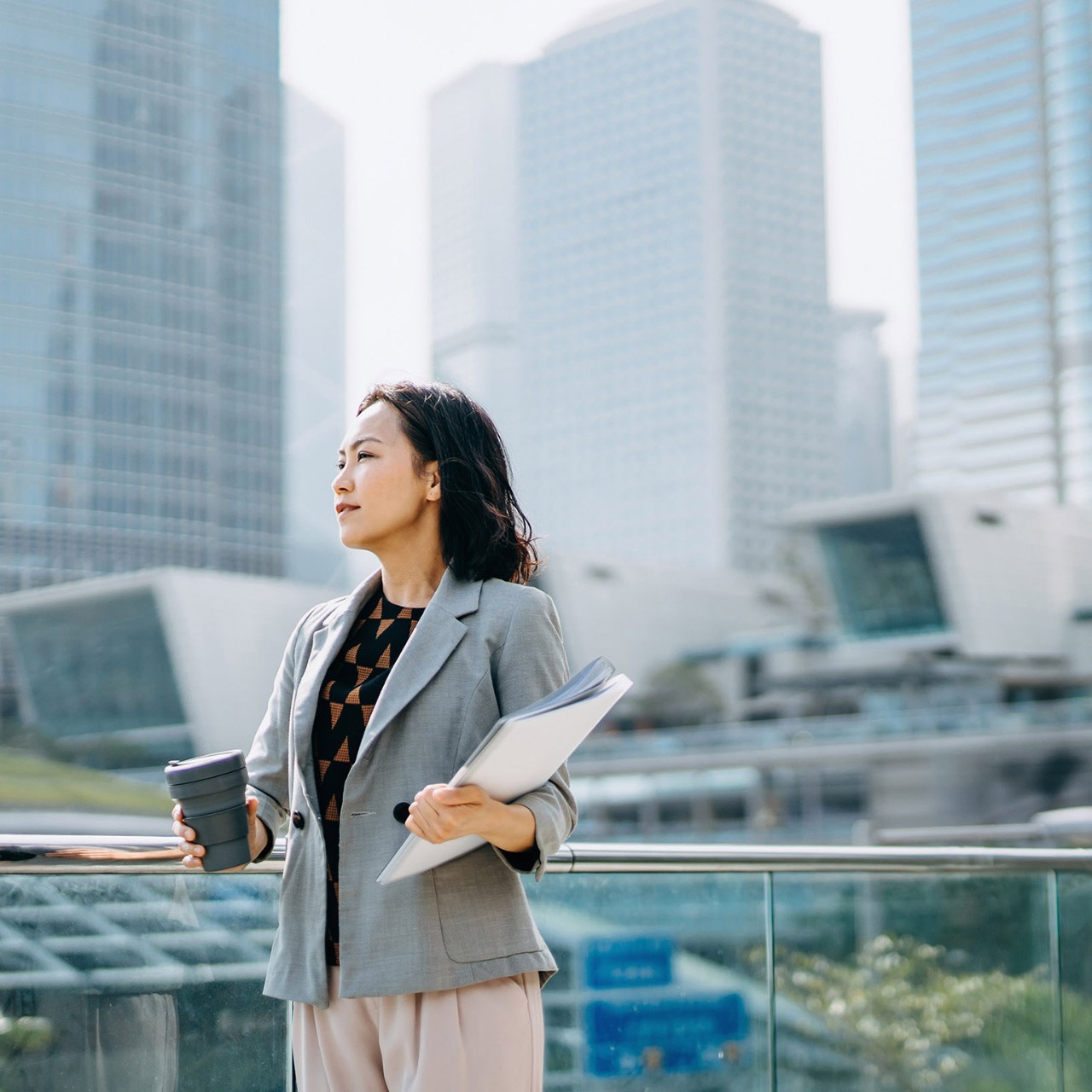 Portrait of confidence Asian businesswoman looking away and holding documents and having coffee to go against city scene in front of modern office buildings