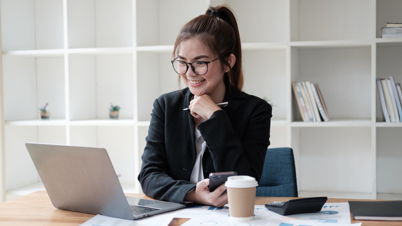 Smart Asian businesswoman working with laptop computer while holding smartphone in her hands, sitting at office desk.