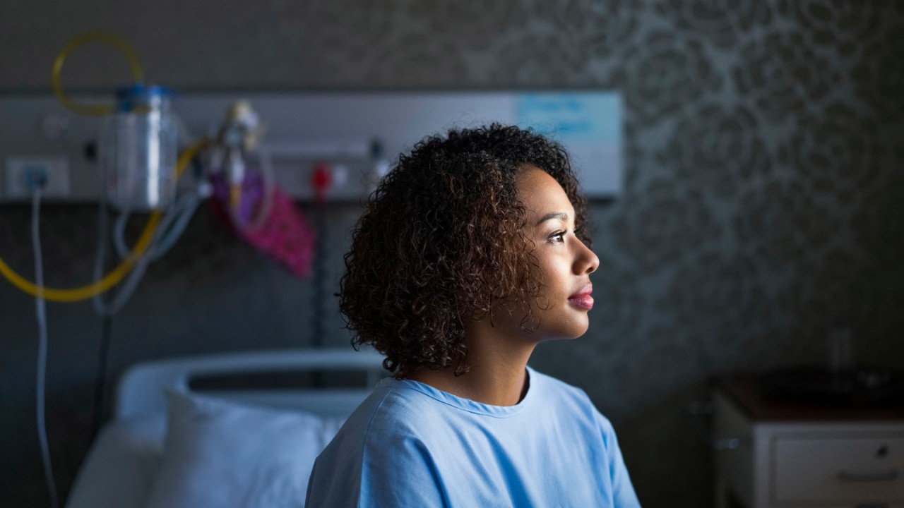 Young woman with curly hair sitting at hospital. Female patient is on bed in ward. She is wearing blue gown.