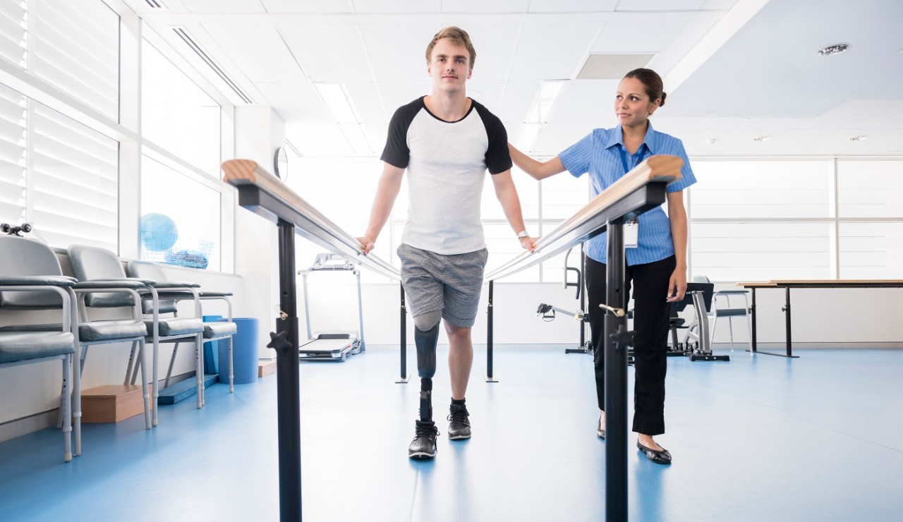 Female physiotherapy helping young male amputee to walk. Patient wearing prosthesis using rehabilitation equipment. Man learning to walk again. Rehabilitation, recovery, determination, physiotherapy.