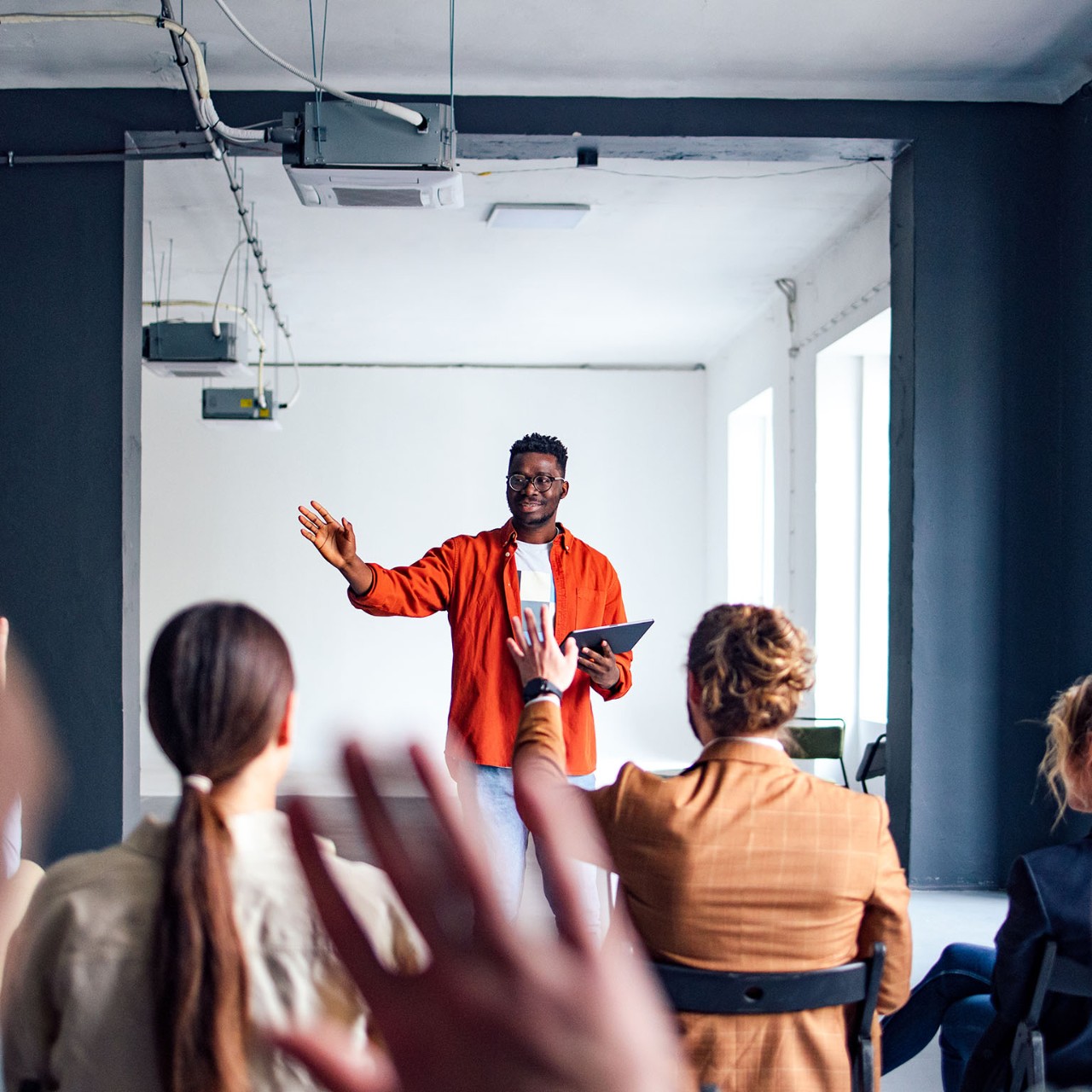 Handsome cheerful man in a orange shirt standing in front of an audience holding a tablet and using hand gestures to interact with the audience.