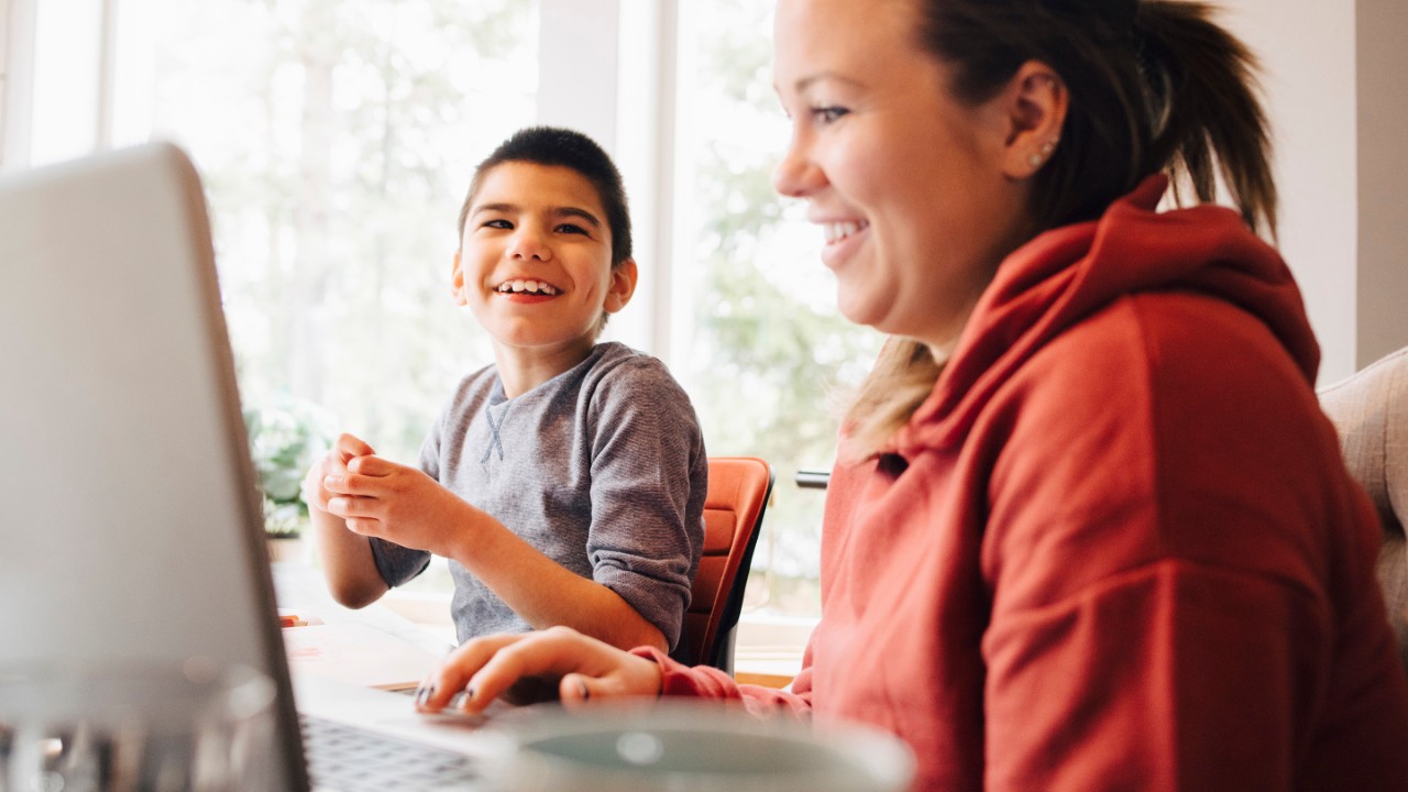 Woman in casual clothes using a laptop while her son smiles next to her at a table.